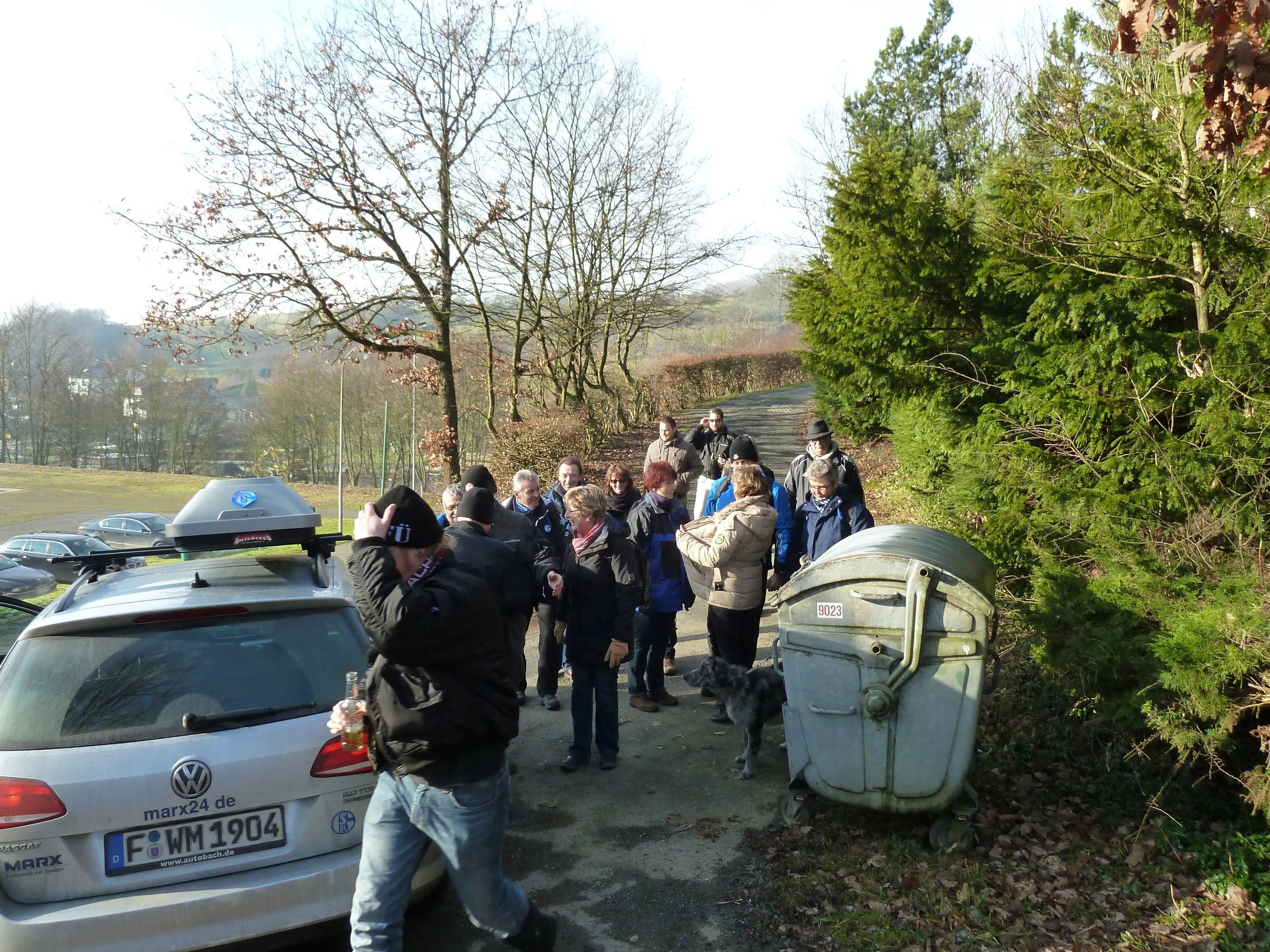 Eine Gruppe von Menschen steht auf einem Weg in einem Park, umgeben von Bäumen. Es ist vermutlich Herbst, da die meisten Bäume keine Blätter haben und einige noch braune Blätter tragen. Einige Leute unterhalten sich, andere fotografieren oder beobach