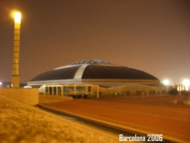 Ein modernes gebäudeartiges Bauwerk bei Nacht mit einer riesigen, geschwungenen Dachstruktur, umgeben von einer Straße und einer Industrierohrleitung, am unteren Rand steht der Text 'Barcelona 2006'.