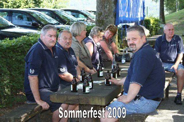 Gruppe von Menschen sitzt an einem Holzbank-Tisch im Freien, es gibt Bierflaschen auf dem Tisch; Text: "Sommerfest 2009"