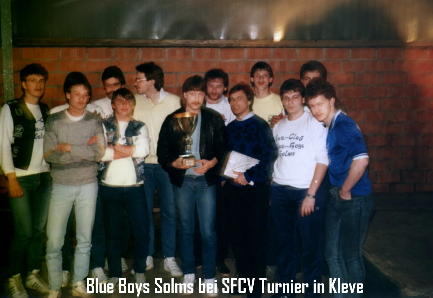 Gruppe von jungen Männern, die einen Pokal bei einem Turnier in Kleve halten, mit einem Backsteinwand im Hintergrund.