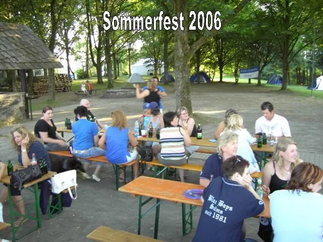 Menschen sitzen an Tischen beim Sommerfest 2006 im Freien, einige trinken Bier, im Hintergrund Zelte und Bäume.