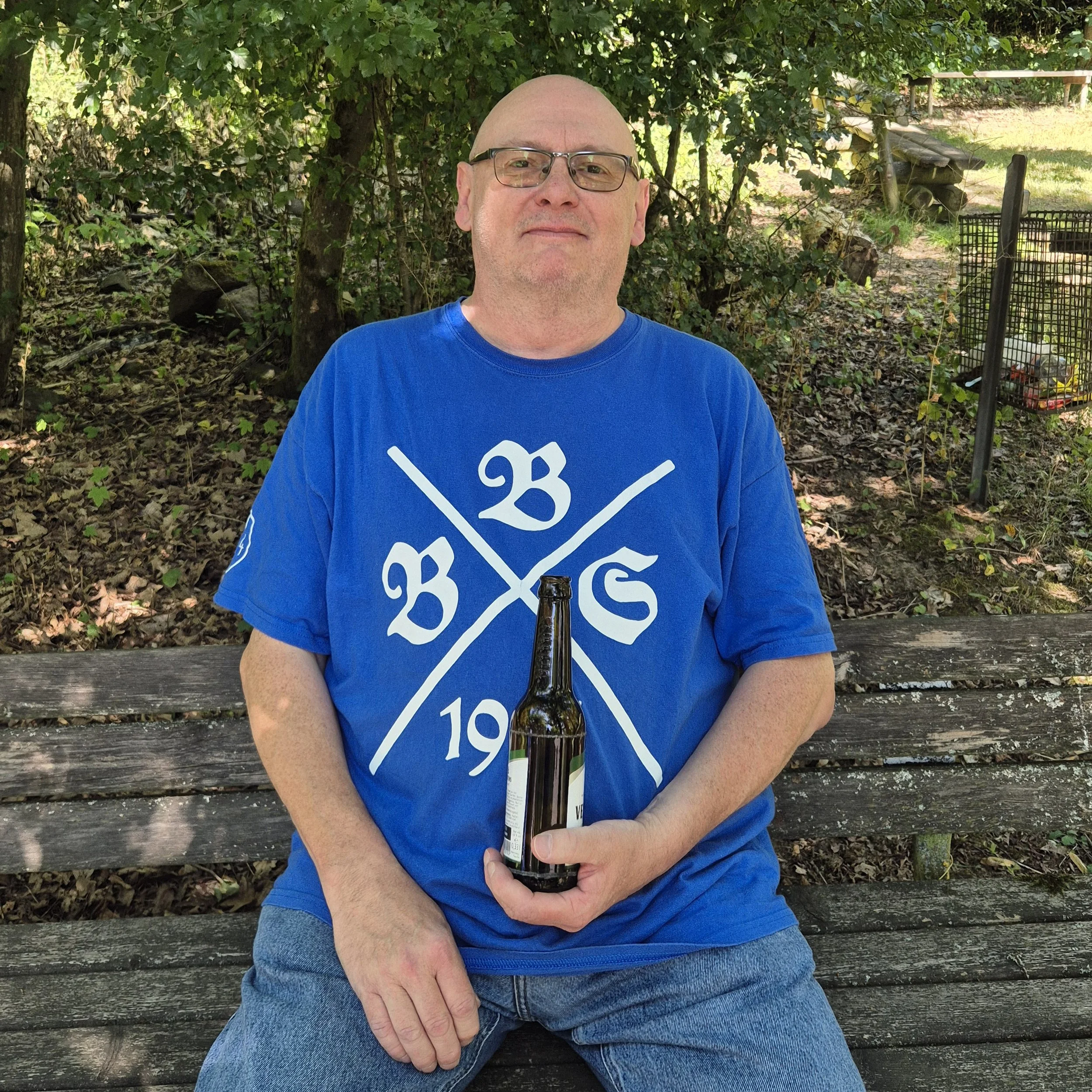 Ein Mann mit Brille sitzt auf einer Bank im Grünen und hält eine Bierflasche in der Hand.
