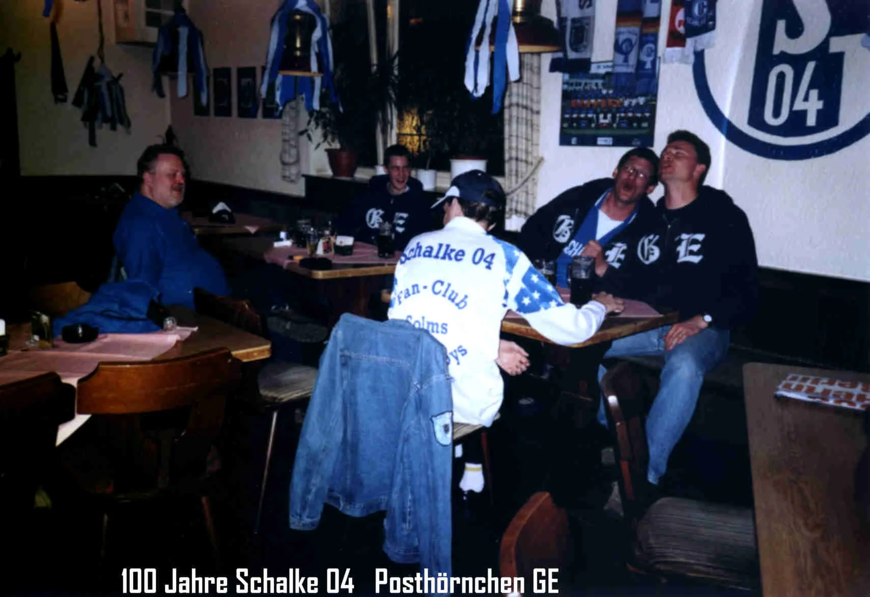 Gruppe von Menschen, die in einer Kneipe unterwegs sind, trägt Schalke 04 Fanartikel, sitzt an Tischen, lacht und unterhält sich. Hintergrund mit Schalke 04 Symbolen und Bannern.