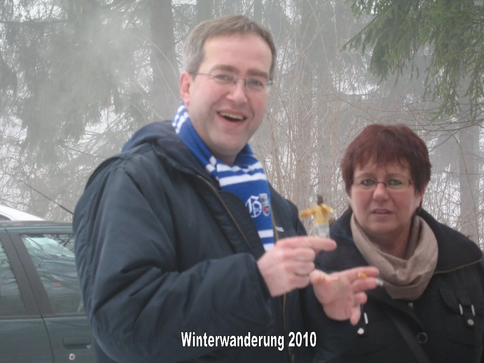 Zwei Menschen, ein Mann und eine Frau, beim Winterwandern im Jahr 2010. Der Mann trägt eine dunkle Jacke und ein blau-weiß gestreiftes Halstuch, während die Frau eine dunkle Jacke und ein helles Schal trägt. Im Hintergrund sind Bäume und ein Auto sic
