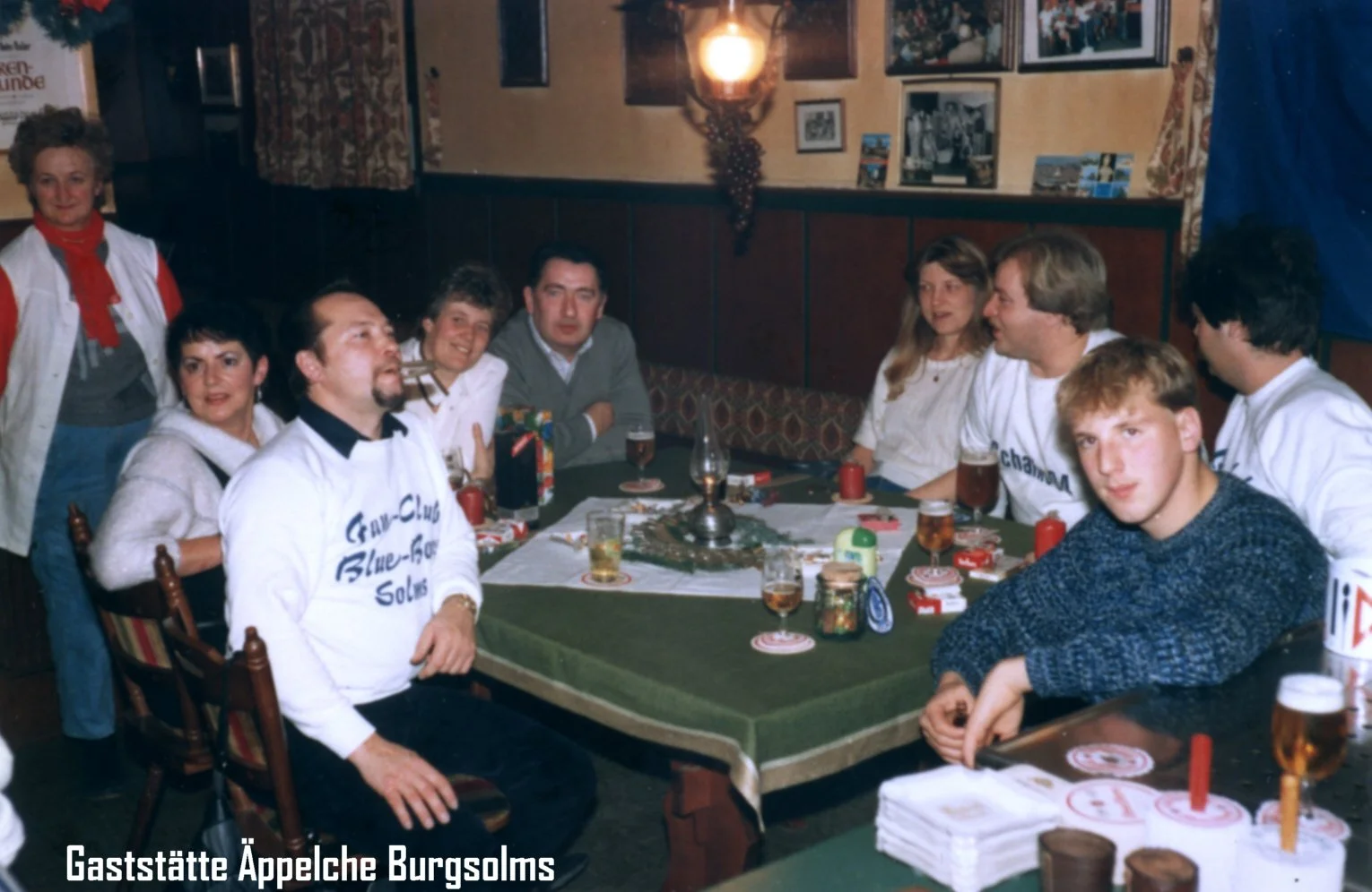Eine Gruppe von Menschen sitzt um einen Tisch in einem gemütlichen Raum, vermutlich in einer Gaststätte. Es gibt Getränke, Kerzen und Staubcoaster auf dem Tisch, die Wände sind mit Fotos dekoriert, und es gibt eine Frau, die links im Bild steht.