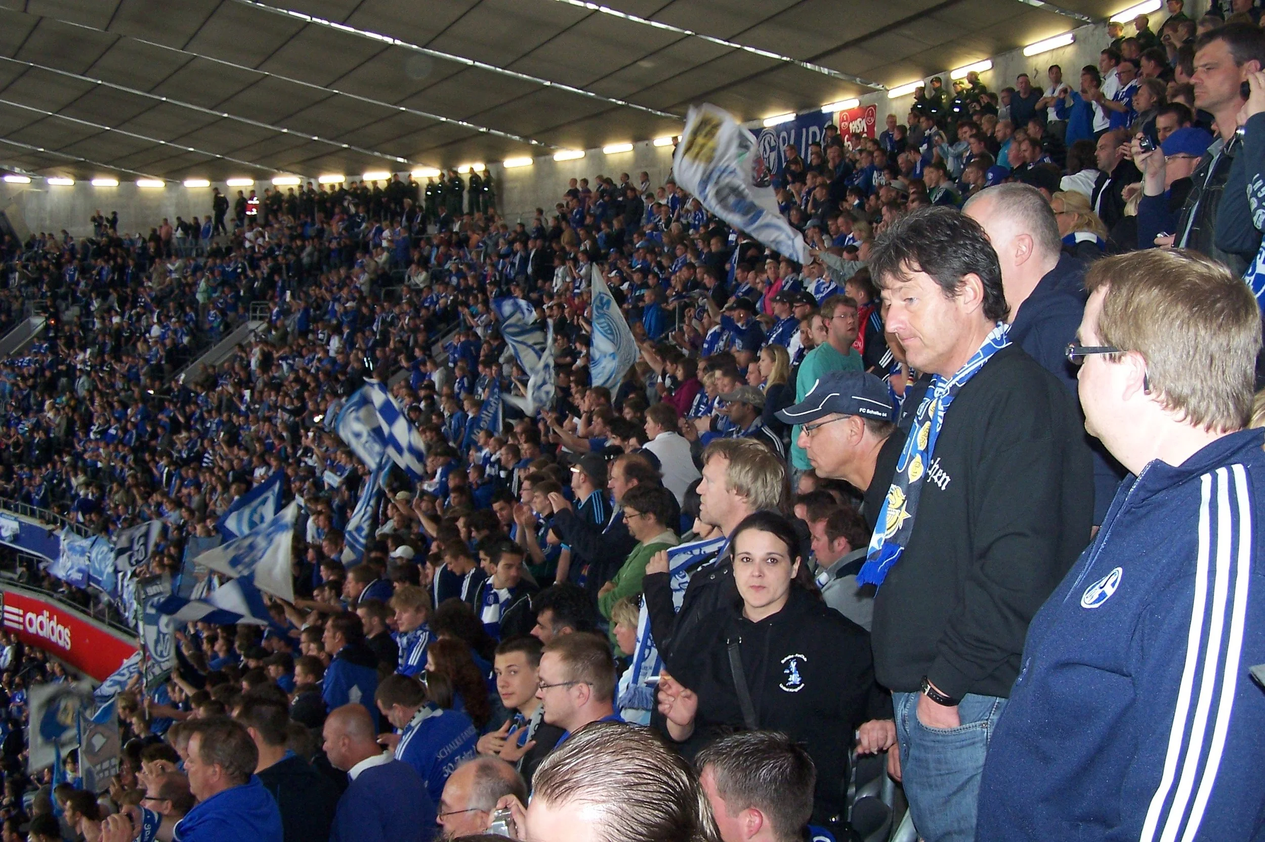 Fußballfans in einem Stadion, die in blauen und weißen Trikots und Schals auf der Tribüne sitzen, mit Flaggen und Bannern.