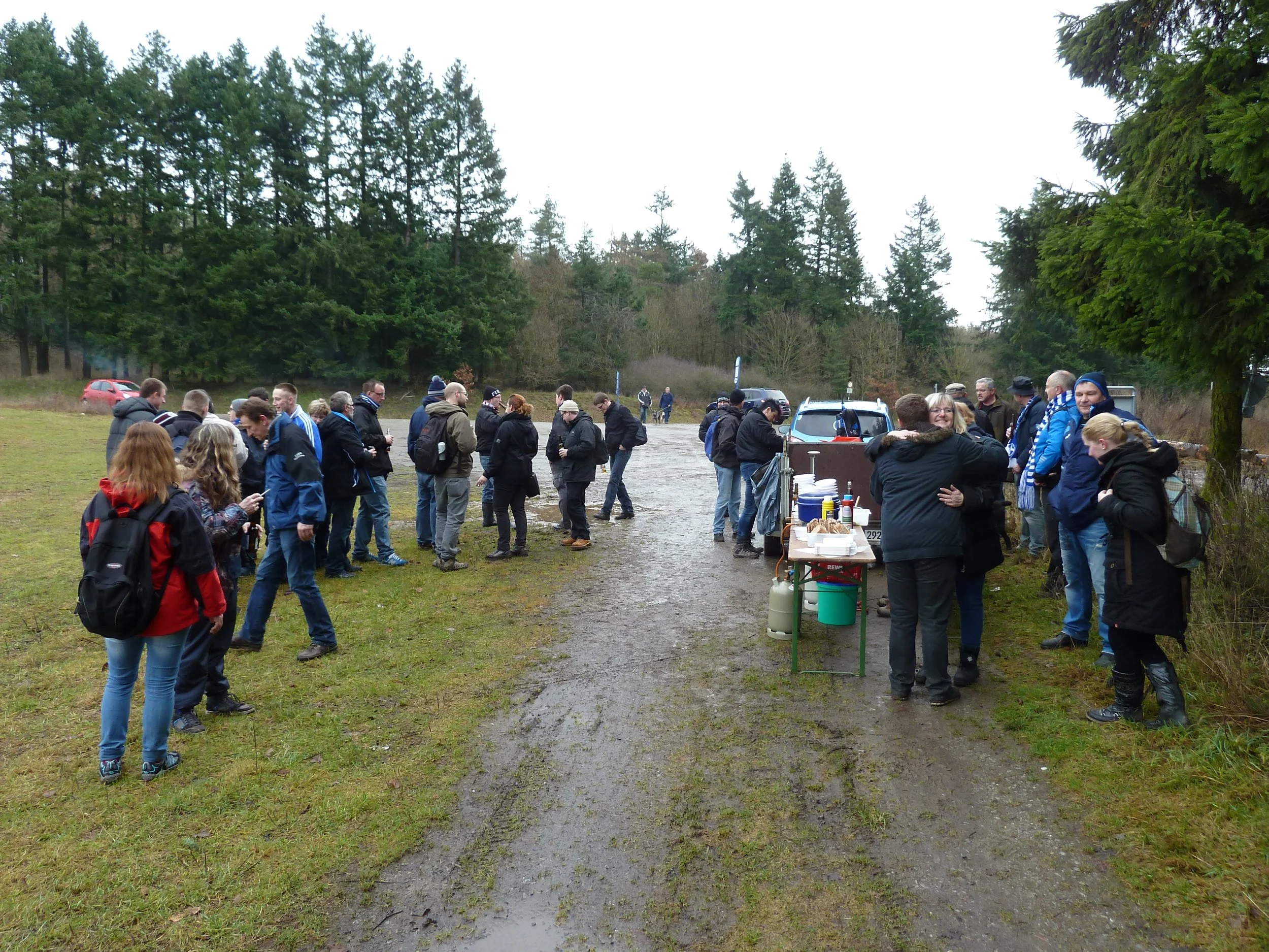 Ein Gruppen von Menschen steht in einer Reihe im Freien bei einem Treffen oder Event, mit Tischen, Getränken und Essen, umgeben von Bäumen und einer offenen Landschaft.