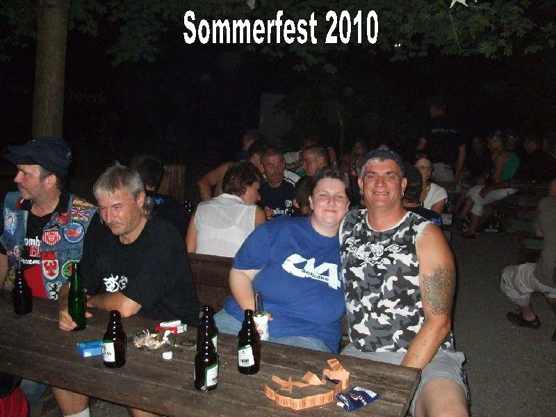 Menschen sitzen bei einem Sommerfest 2010 am Abend im Freien. Zwei Personen stehen im Vordergrund, lachen und haben Getränke vor sich. Es gibt mehr Leute im Hintergrund, bei dunklem Himmel und Bäumen.