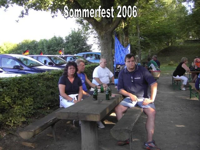 Menschen sitzen an einem Holztisch bei einem Sommertest 2006 im Freien, mit Bäumen und Fahrzeugen im Hintergrund.