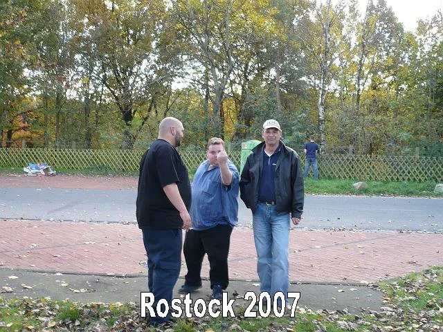 Drei Männer stehen auf einem Bürgersteig im Freien, im Hintergrund Bäume mit Herbstlaub, Rostock 2007.