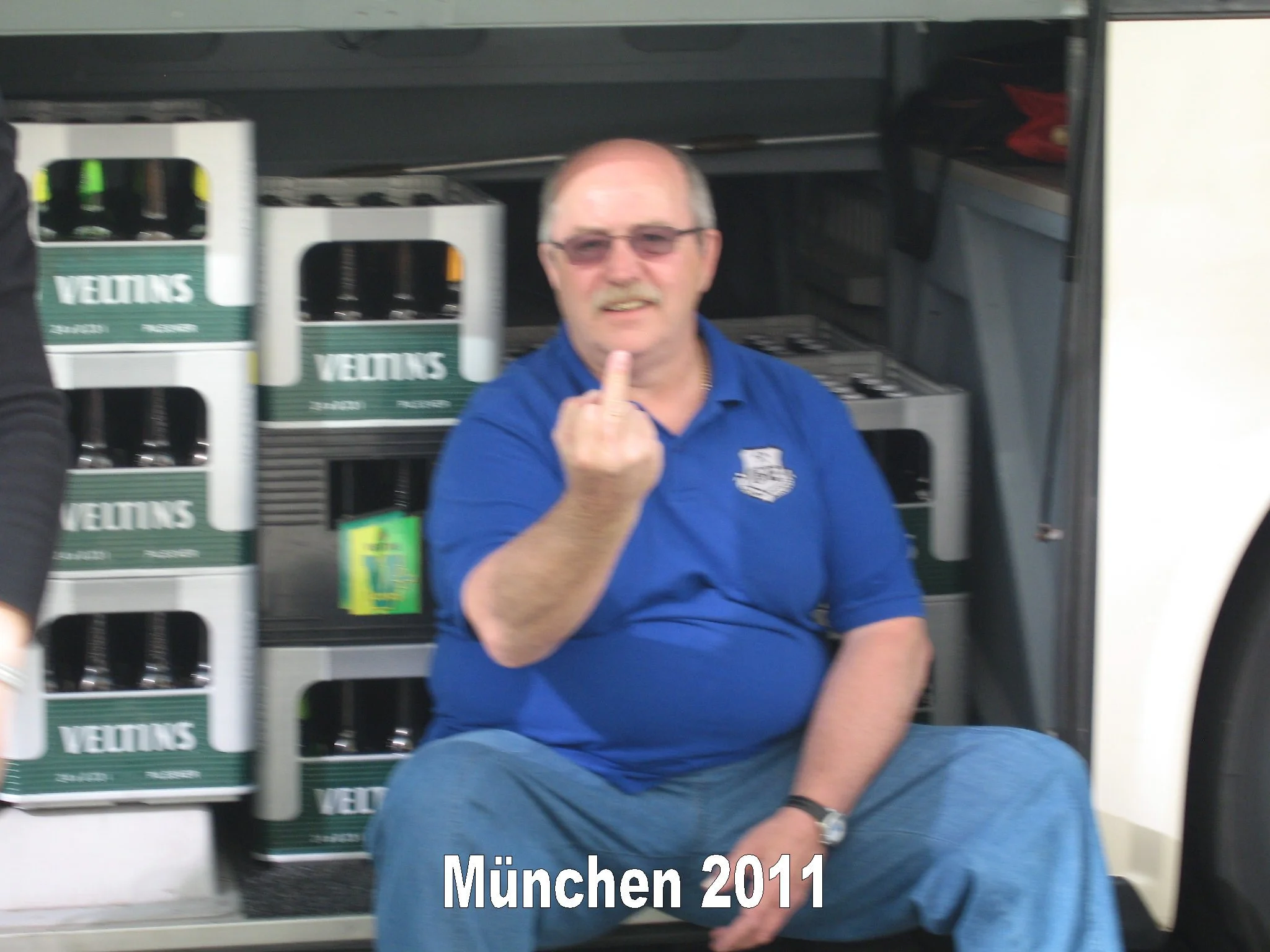 Ein Mann trägt eine blaue Polo-Hemd und sitzt vor einem Regal mit Bierkästen. Im Vordergrund steht der Text 'München 2011'.