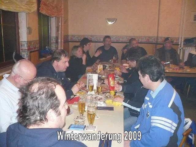 Gruppe von Menschen sitzt an einem langen Tisch in einer Gaststätte, trinkt Bier und unterhält sich. Auf dem Tisch sind Getränke, Servietten und Flaschen. Der Raum hat eine warme Beleuchtung und Fenster an der Seite. Es scheint eine gesellige Zusamme