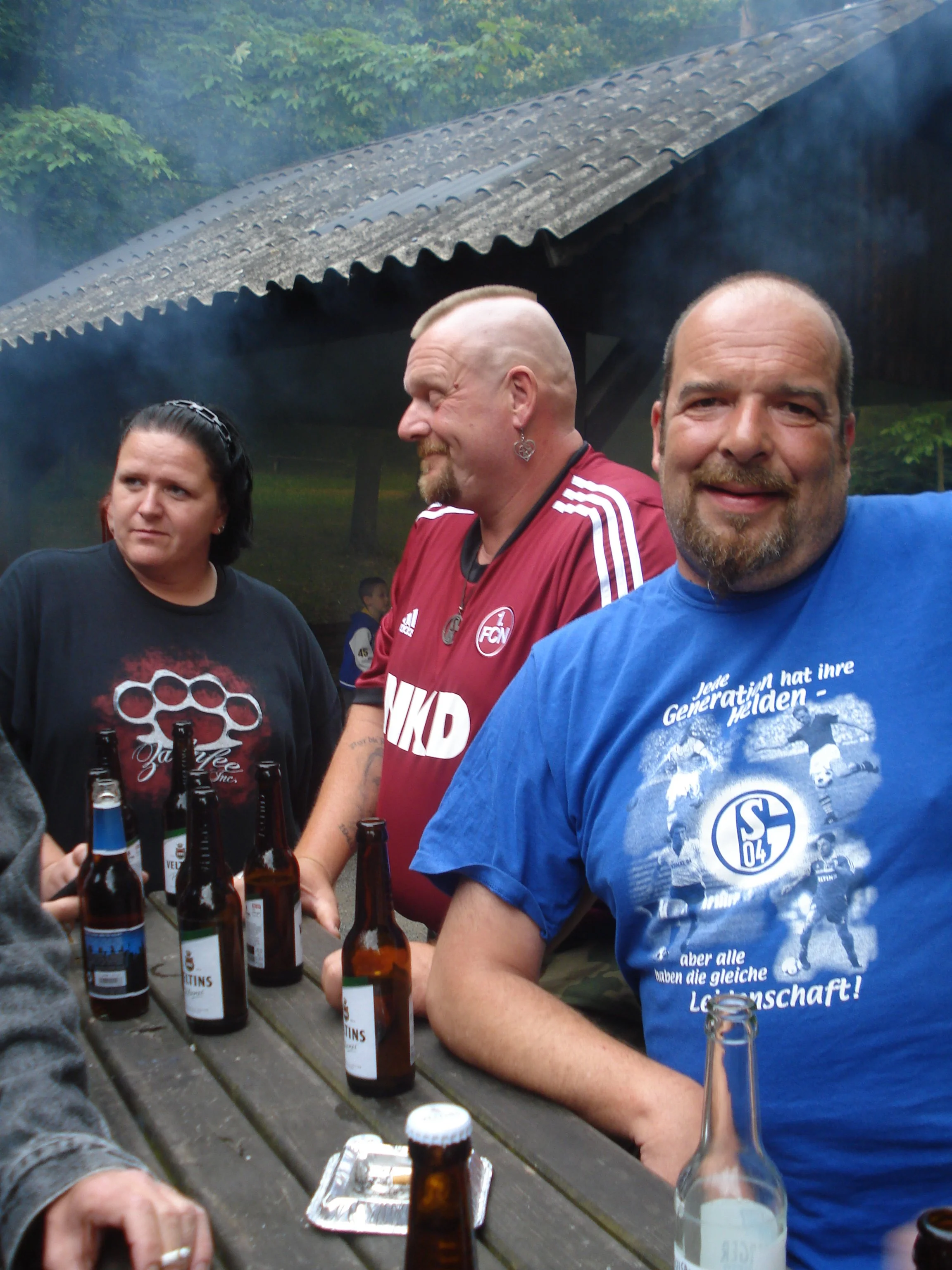 Gruppe von Menschen beim Treffen mit Bierflaschen auf einem Holztisch im Freien, mit Bäumen im Hintergrund.