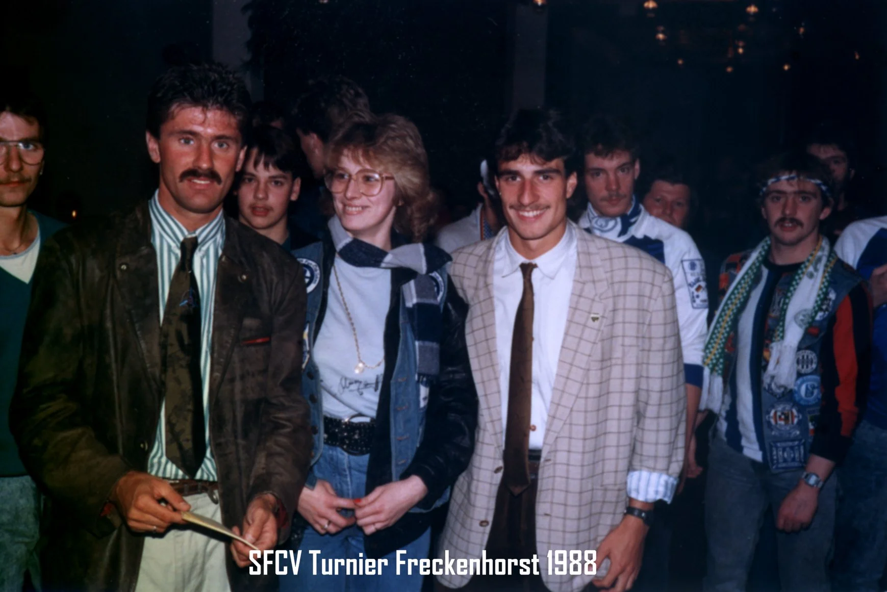 Eine Gruppe von Menschen, darunter Männer und Frauen, beim Turnier in Freckenhorst 1988. Einige tragen Sportbekleidung, andere formelle Kleidung oder typische Mode der 1980er Jahre. Im Vordergrund sind drei Männer und eine Frau zu sehen, die freundli
