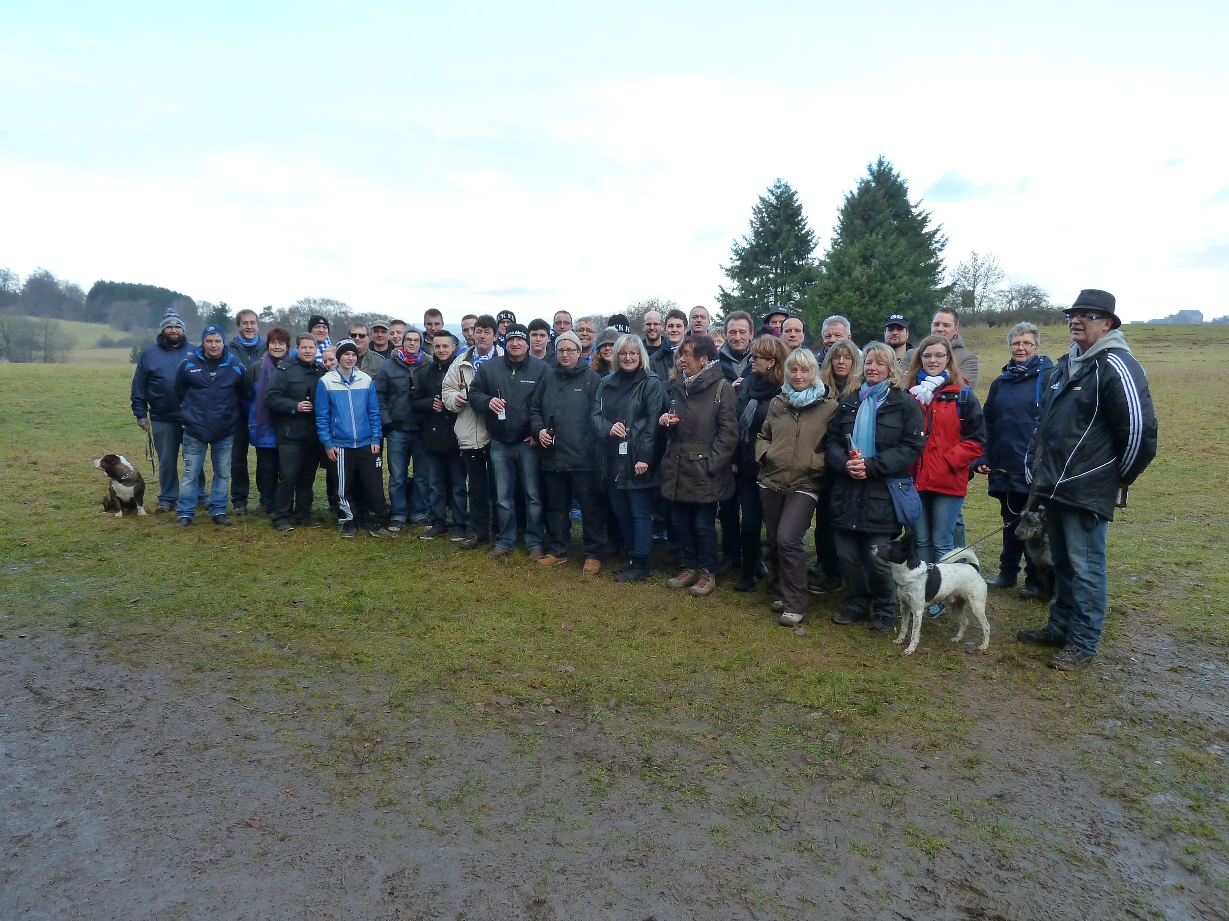 Gruppenfoto von Menschen, die im Freien auf einer Wiese stehen, viele tragen Jacken und Mützen, einige halten Getränke, ein Hund ist sichtbar, hinter ihnen Bäume und ein bewölkter Himmel.