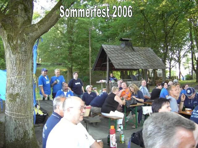 Gruppen von Menschen sitzen an Tischen im Freien beim Sommerfest 2006, unter Bäumen mit einem kleinen Haus im Hintergrund.