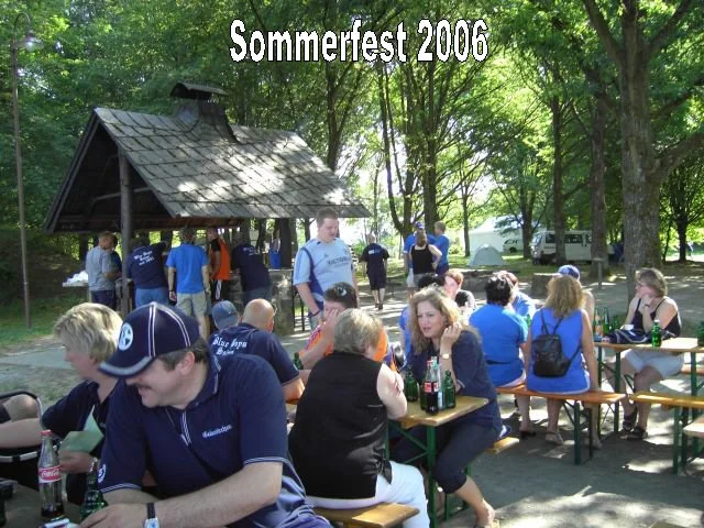 Menschen sitzen an Tischen beim Sommerfest 2006 unter Bäumen, im Hintergrund ein Holzunterstand.