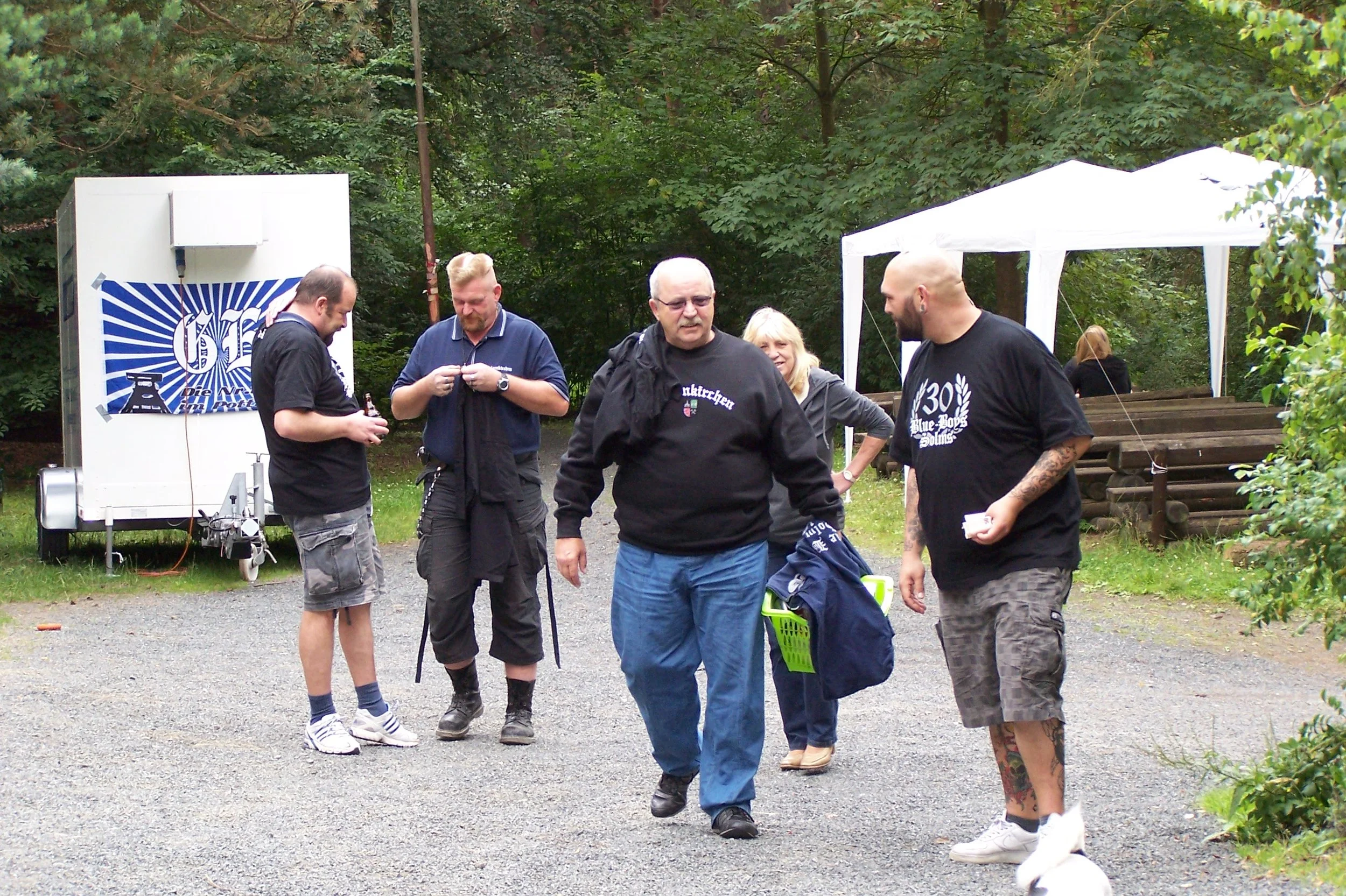 Gruppe von Menschen im Freien, wahrscheinlich bei einer Veranstaltung, mit Bäumen im Hintergrund, einige sprechen miteinander.