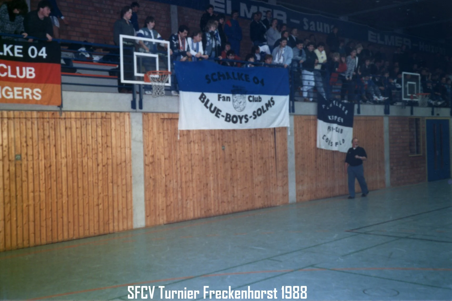 Innenansicht eines Sporthallens mit Vereinskämmen und Menschen auf der Tribüne. Ein Handballkorb ist sichtbar. Auf der Tribüne hängen Banner, darunter eines mit der Aufschrift "Schalke 04" und "Blue-Boys-Solms". Am unteren Rand steht der Text "SFCV T