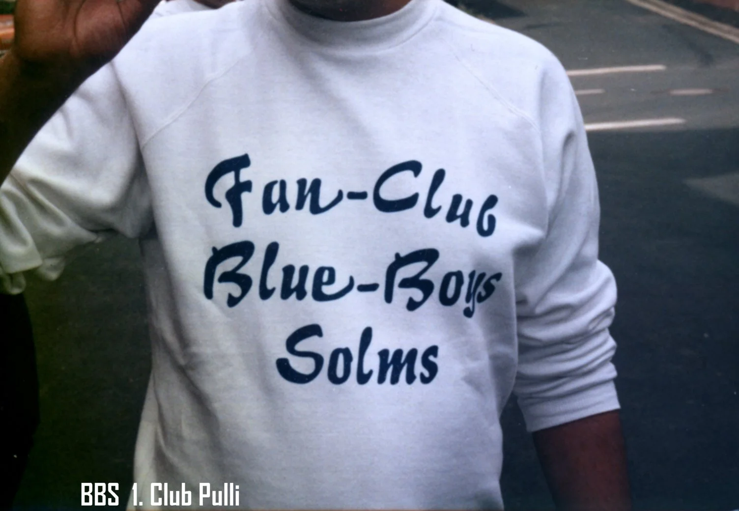 Person trägt einen weißen Pullover mit blauer Schrift, die sagt: 'Fan-Club Blue-Boys Solms'.