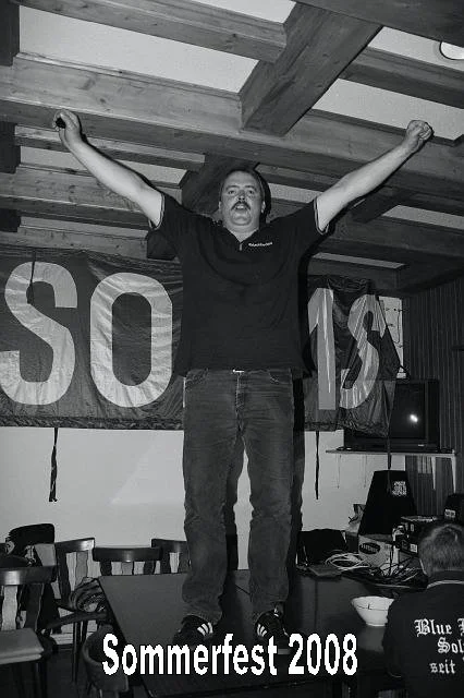 Mann steht auf Tisch mit ausgestreckten Armen bei Sommerfest 2008, schwarz-weiß Foto, im Hintergrund Holzdecke, Banner im Hintergrund, Küche mit Fernseher und Handys auf Tischen