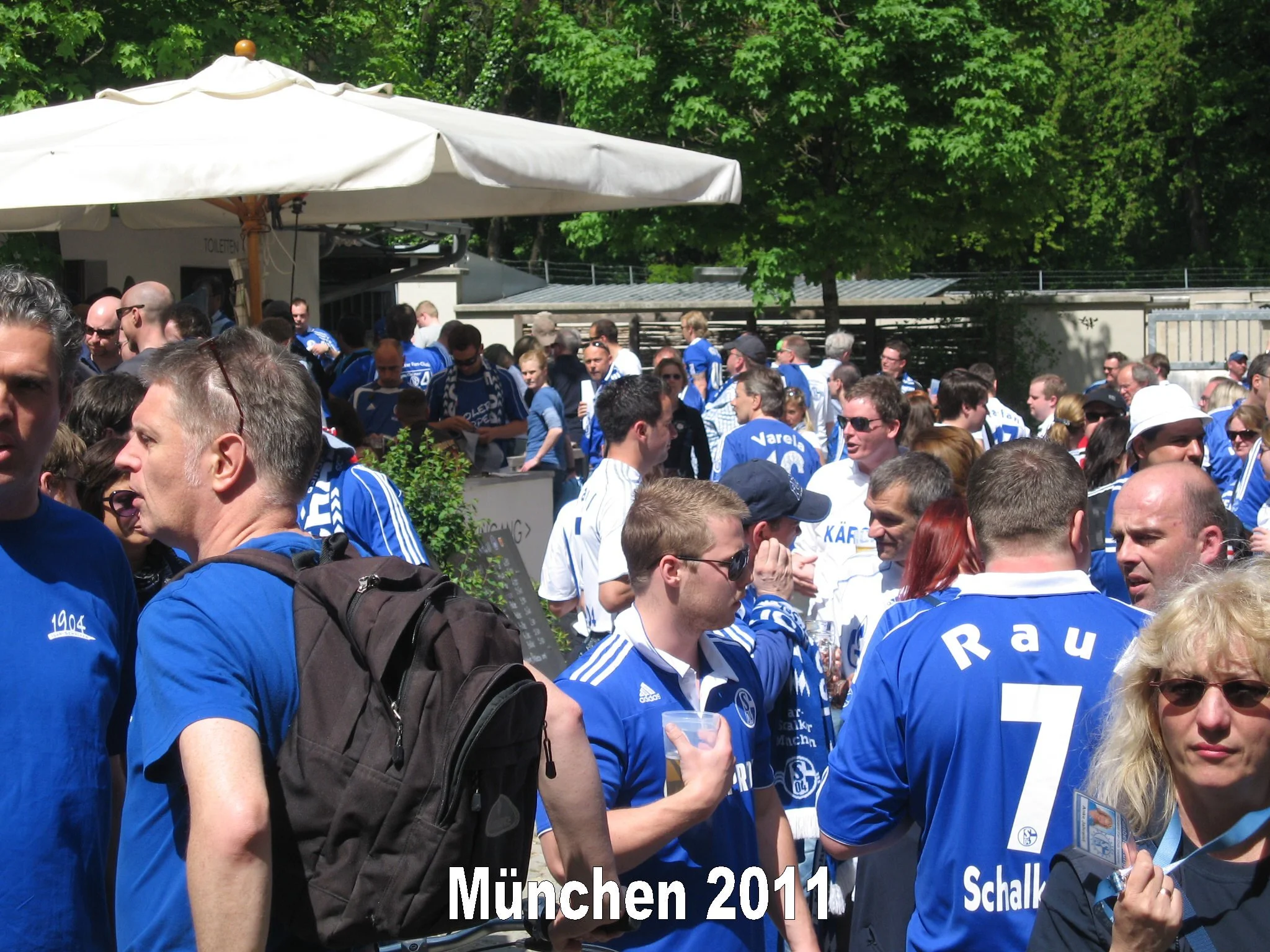 Menschenmenge bei einem Sportereignis in München 2014, viele tragen blaue Trikots und Hoodies, es gibt Bäume im Hintergrund und einen Sonnenschirm, einige tragen Sonnenbrillen.