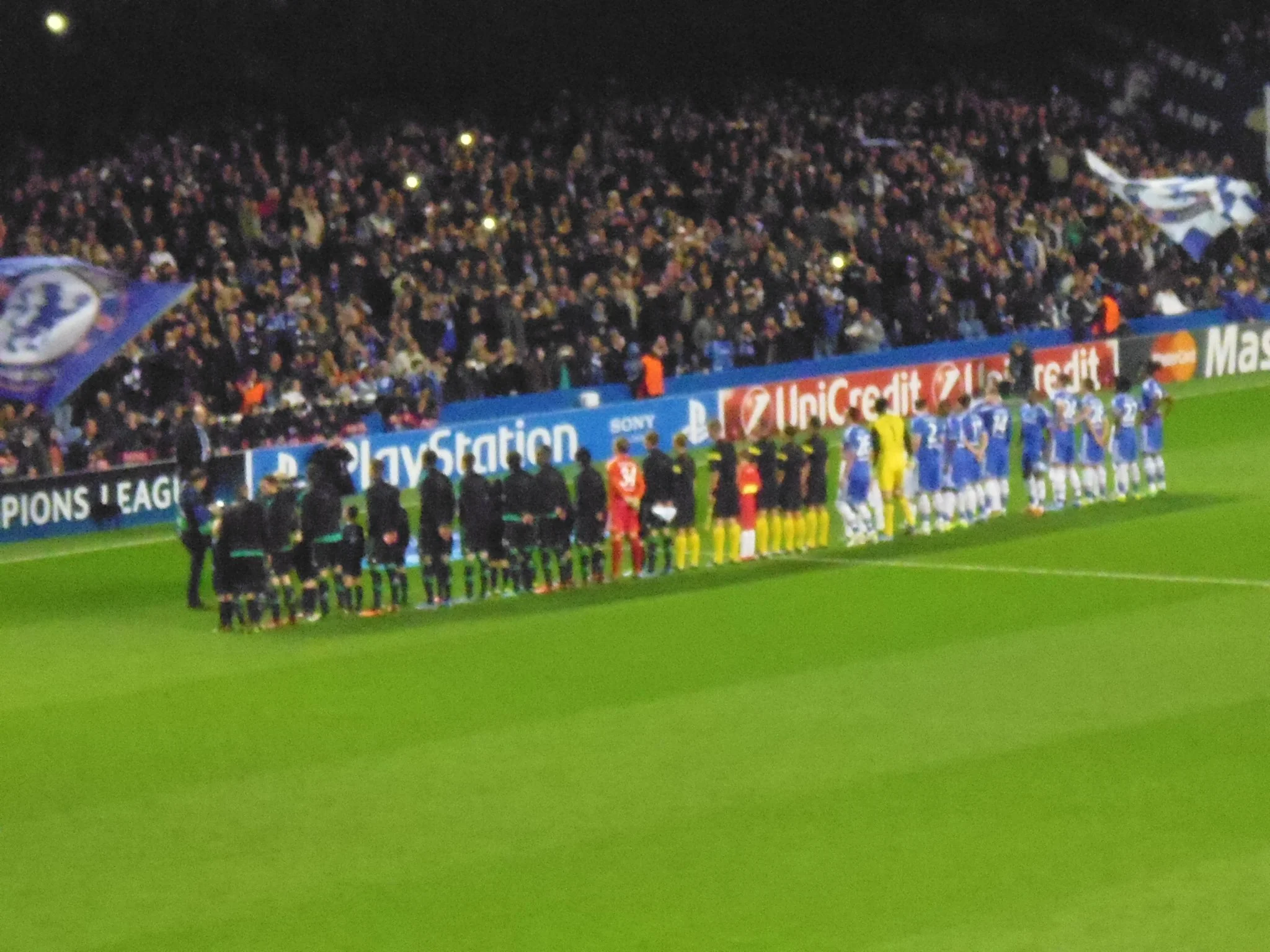 Fußballspiel zwischen zwei Mannschaften auf einem Stadion. Die Spieler stehen in einer Reihe, während die Zuschauer im Stadion den Spielverlauf beobachten.