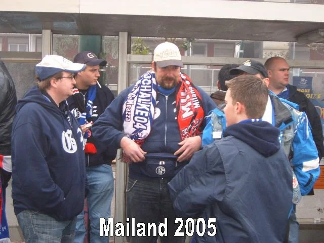 Gruppe von jungen Männern, die sich unter einer Brücke versammelt haben. Einige tragen Schal- und Kapuzenjacken, vermutlich Fußballfans. Es ist regnerisch, im Hintergrund gehen Menschen vorbei. Text auf dem Bild: "Mailand 2005"