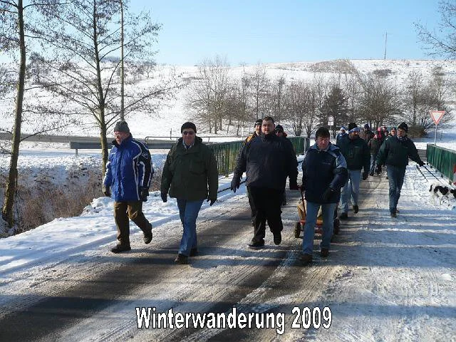 Menschen ziehen während einer Winterwanderung 2009 auf einem verschneiten Weg, mit Bäumen und Hügel im Hintergrund.