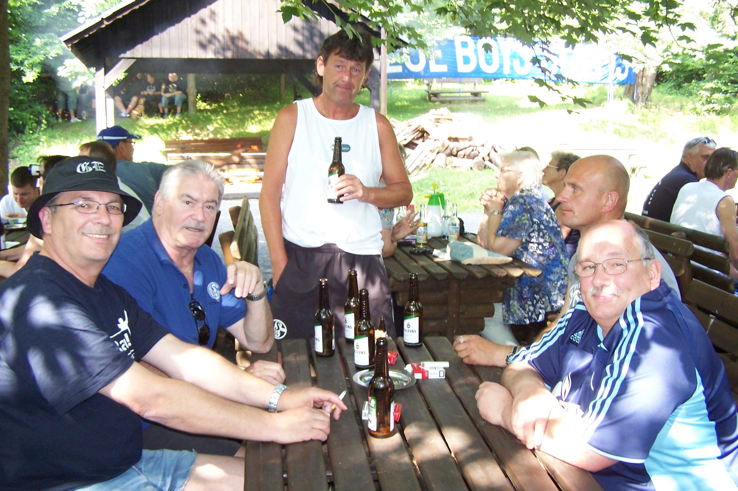 Gruppe von Menschen, die im Freien an einem Picknick oder Grillfest teilnehmen, mit Getränken und Essen auf Holztischen, unter Bäumen im Hintergrund.