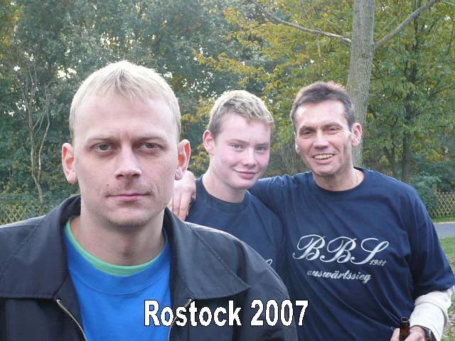 Drei Männer stehen im Freien, im Hintergrund Bäume, im Vordergrund ein Mann mit blondem Haar, der ernst schaut, und im Hintergrund ein Mann, der lächelnd und mit einem anderen jungen Mann lächelnd dasteht, alle tragen dunkle T-Shirts. Text auf dem Bi