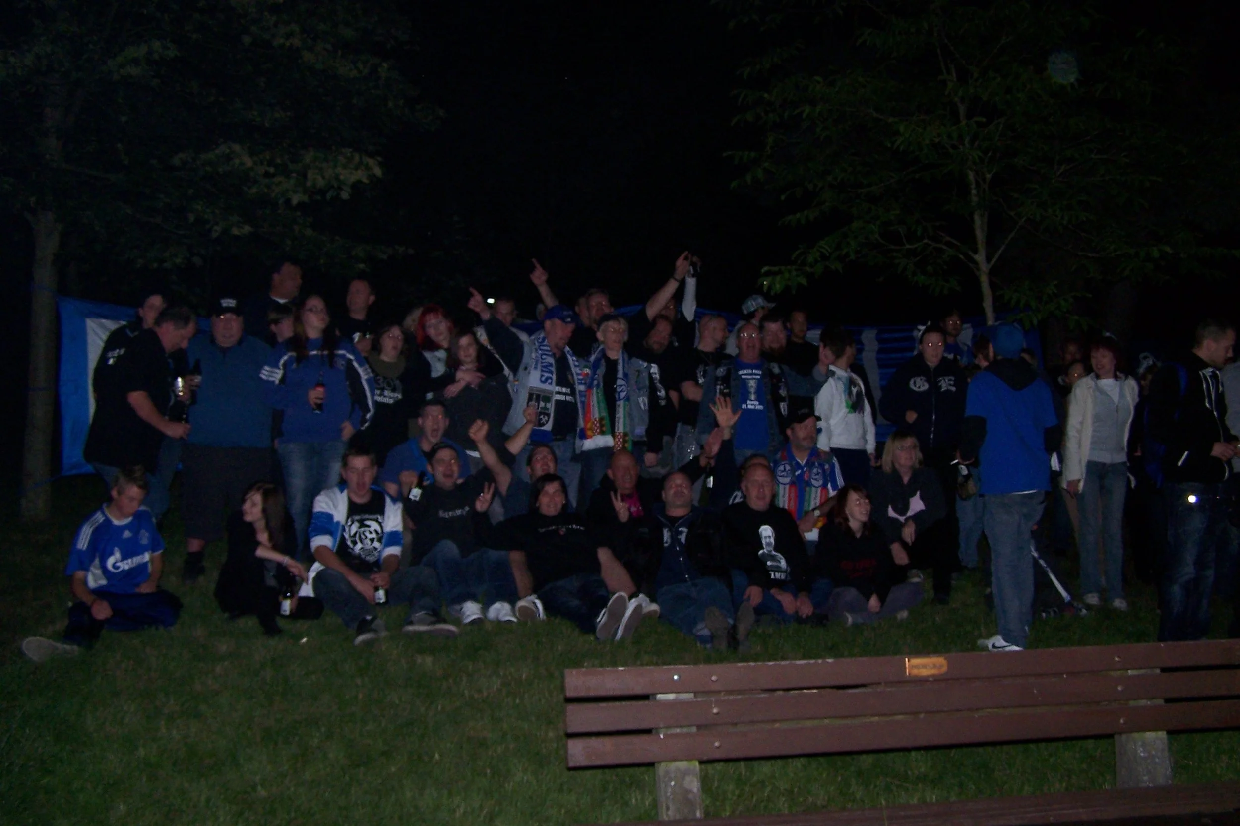 Gruppe von Menschen, die bei Nacht draußen versammelt sind, einige tragen dunkle Kleidung mit blauen Akzenten, und es scheinen Fans eines Sports Teams zu sein, da einige Trikots und Schals sichtbar sind.