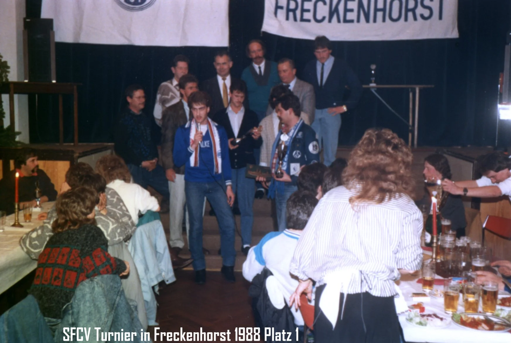 Gruppenfoto bei einem Turnier in Freckenhorst 1988 mit mehreren jungen Männern auf der Bühne, einer hält eine Trophäe, im Vordergrund sitzen mehrere Menschen bei einem Festmahl mit Bier. Im Hintergrund ein Banner mit der Aufschrift 'FRECKENHORST' und