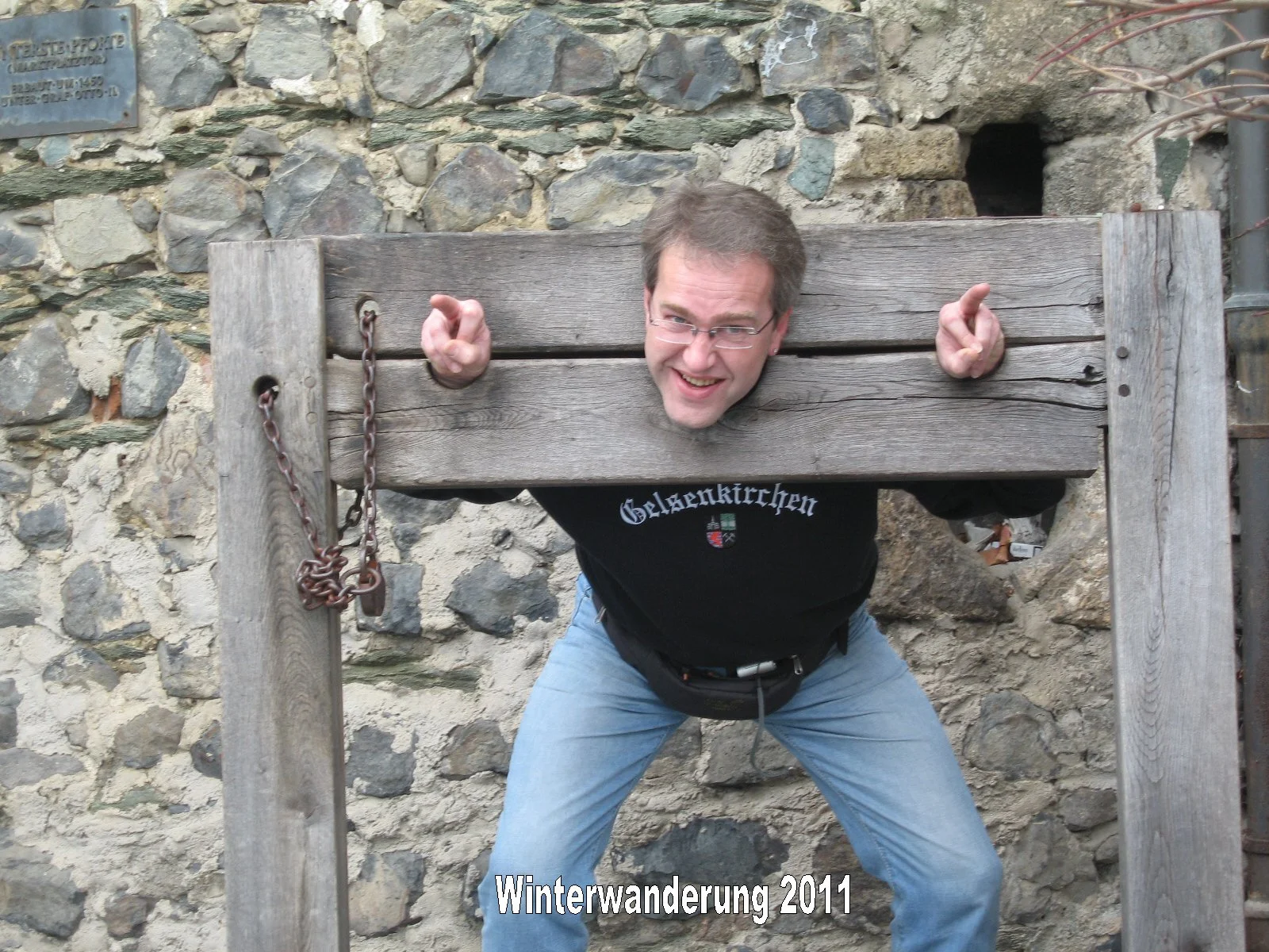 Ein Mann stecken in einem Holzrahmen, der an einer Steinmauer befestigt ist, und macht eine lustige Pose. Er trägt eine Brille und ein schwarzes T-Shirt mit dem Text 'Gelsenkirchen'.