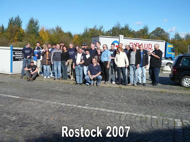 Gruppe von Personen vor einem LKW im Freien, mit dem Text "Rostock 2007" und einem Hintergrund aus Bäumen und einem blauen Himmel.