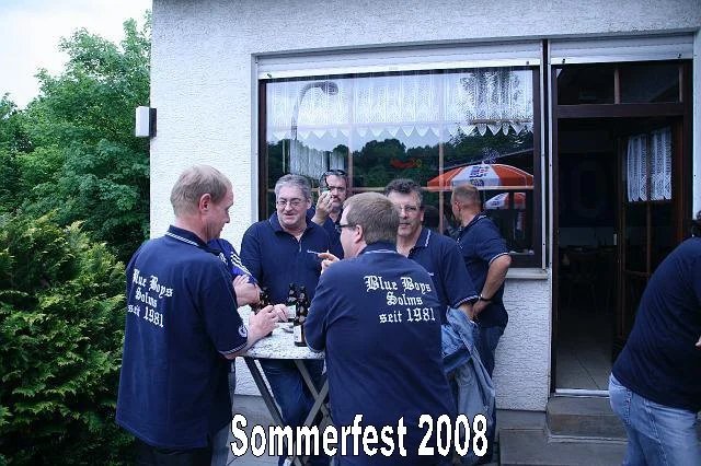 Gruppe von sechs Männern in blauen Shirts, die bei einem Sommerfest 2008 vor einem Haus mit einem Fenster und einer Tür stehen und Getränke trinken.