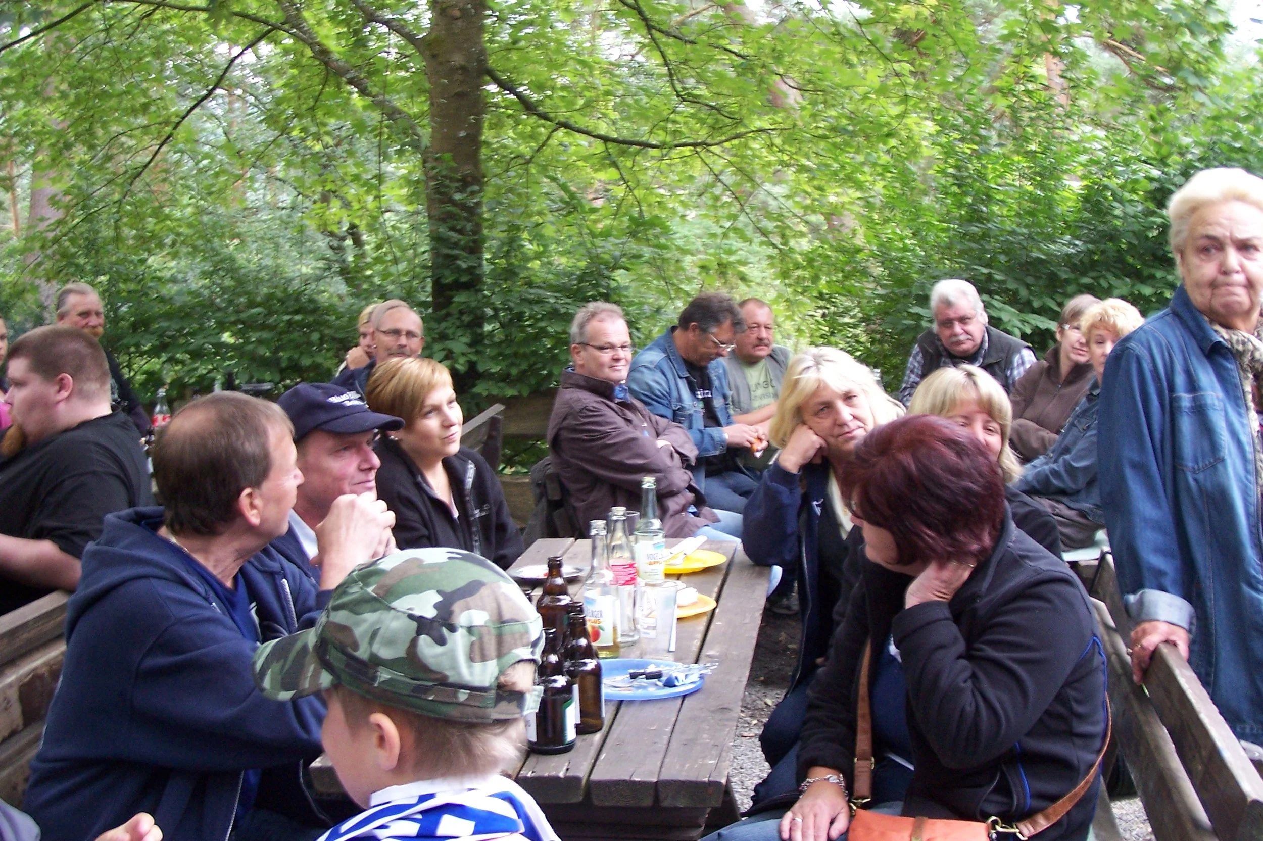 Gruppe von Menschen sitzt im Freien auf Bänken und Essentischen, umgeben von Bäumen, bei einem Treffen oder einer Veranstaltung, mit Getränken und Essen auf den Tischen.