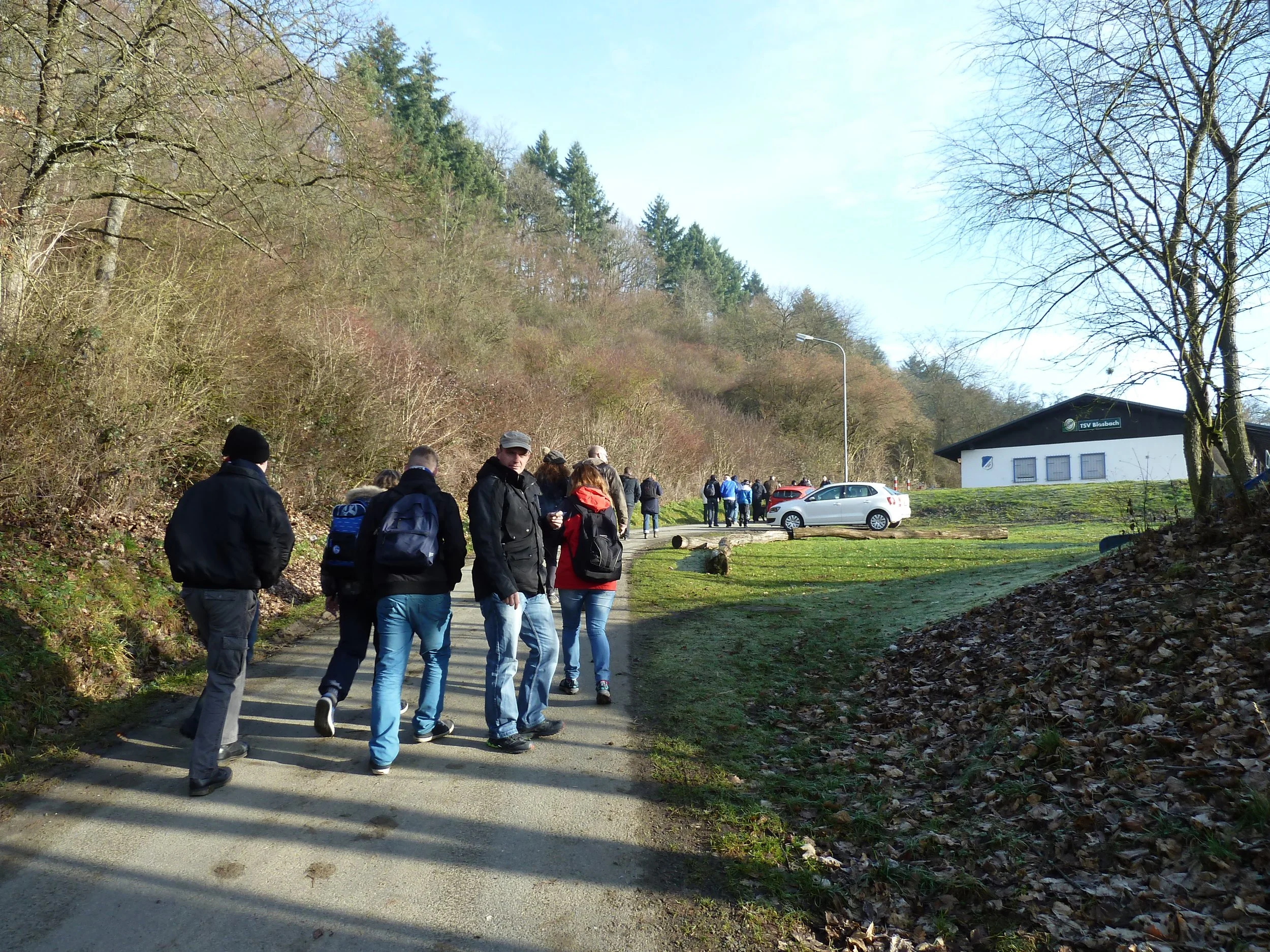 Gruppe von Menschen, die einen Weg in der Natur entlang gehen, umgeben von Bäumen und einem Gebäude im Hintergrund.