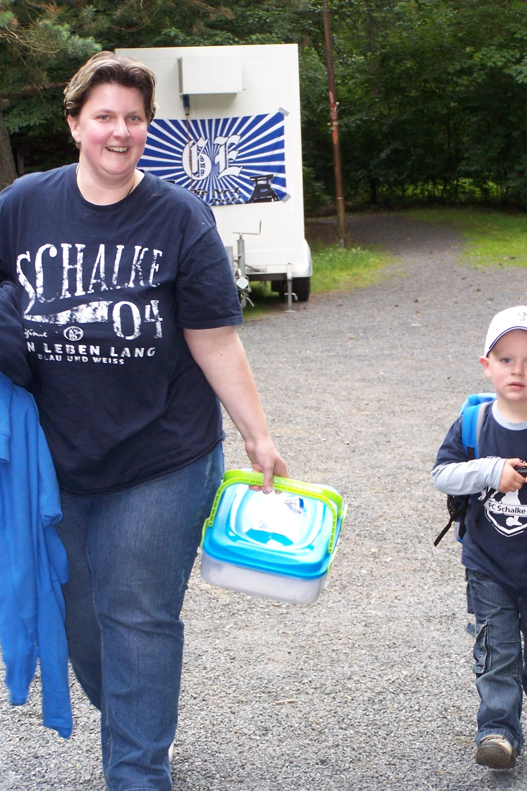 Eine lachende Frau trägt einen blauen Rucksack und eine Kühlbox, im Hintergrund sind grüne Bäume und ein Sommer-Event mit dem Logo von FC Schalke 04 auf einem Banner.