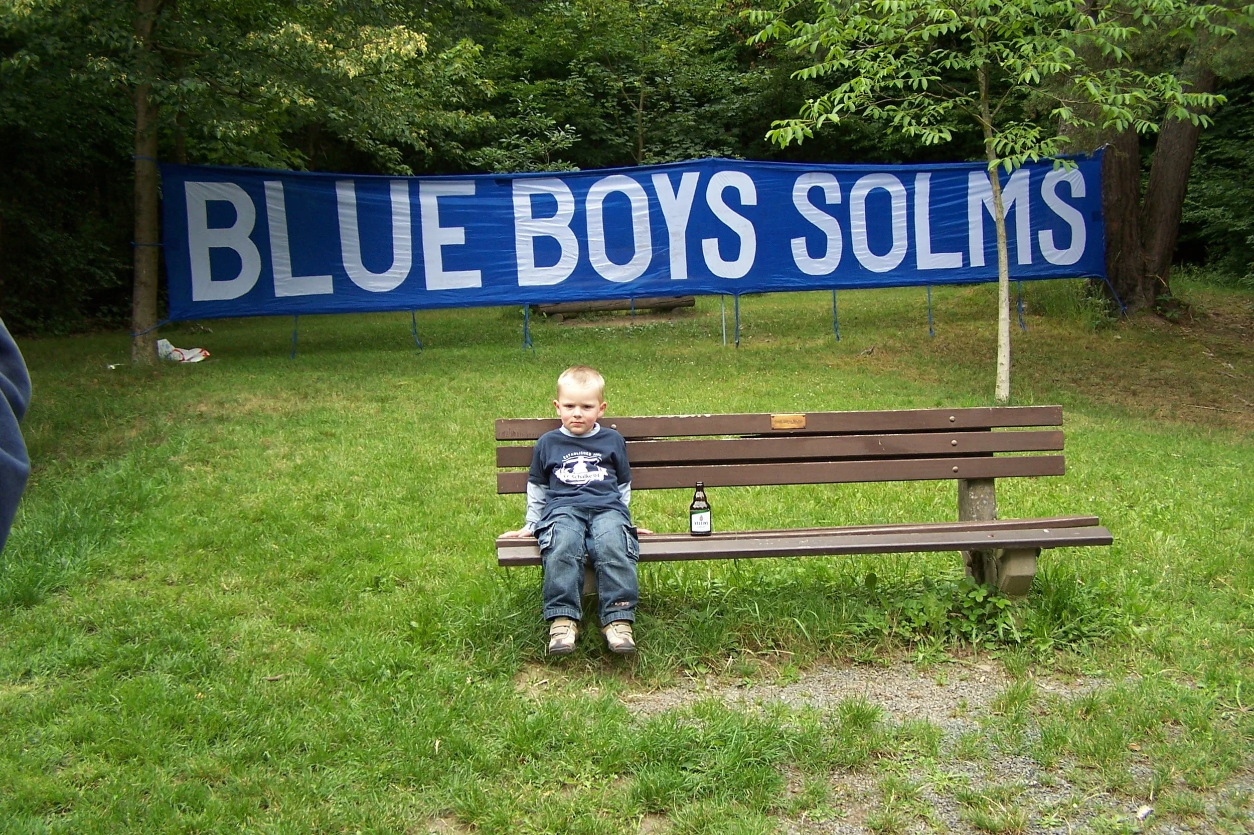 Ein Junge sitzt auf einer Parkbank vor einem blauen Banner mit weißen Buchstaben, die 'BLUE BOSS SULM' sagen, in einer grünen, bewaldeten Umgebung.