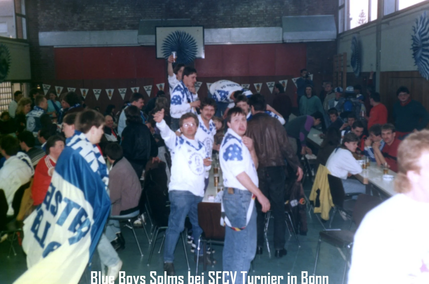 Freudige Menschen im Saal bei einem Turnier, viele tragen blaue und weiße Trikots mit dem Logo des Vereins, einige halten Getränke, im Hintergrund sind Banner und eine Basketballdribbel-Dekoration zu sehen, Deutschland