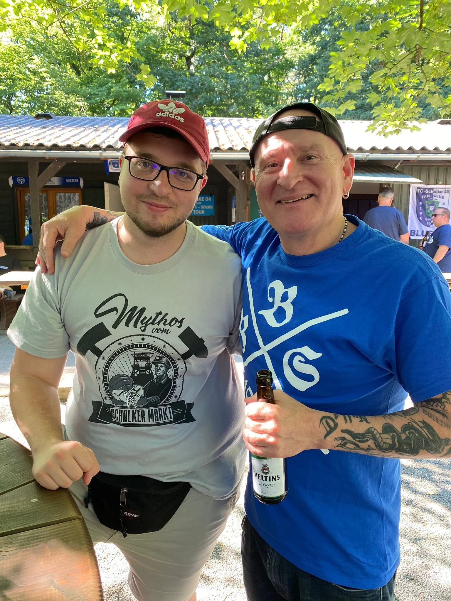 Zwei Männer beim Feiern im Freien, einer hält ein Bier in der Hand. Im Hintergrund sind weitere Personen, Bäume und ein Vereinsschild zu sehen.