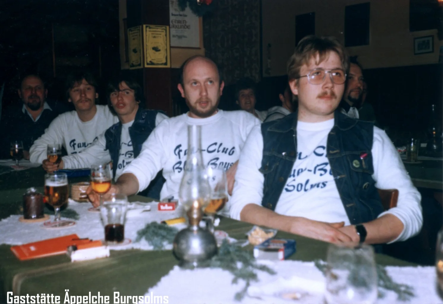 Menschen sitzen an einem Tisch in einer Bar oder einem Restaurant, einige tragen T-Shirts mit dem Text 'Fan-Club Burg Solms'. Es sieht nach einer Feier oder Zusammenkunft aus.