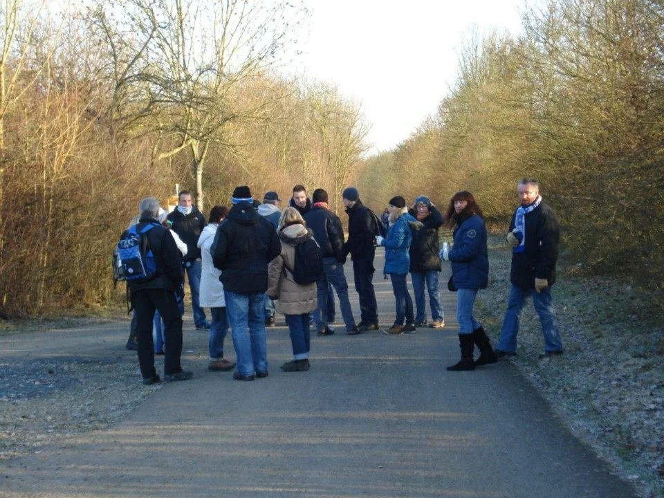 Gruppe von Menschen steht auf einer Straße in einer ländlichen Gegend im Winter, die meisten tragen Jacken und haben Taschen, einige tragen Gesichtsmasken.