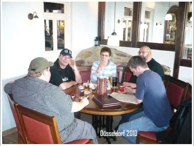 Fünf Personen sitzen um einen runden Tisch in einem gemütlichen Restaurant in Düsseldorf im Jahr 2010. Sie trinken Getränke und unterhalten sich.