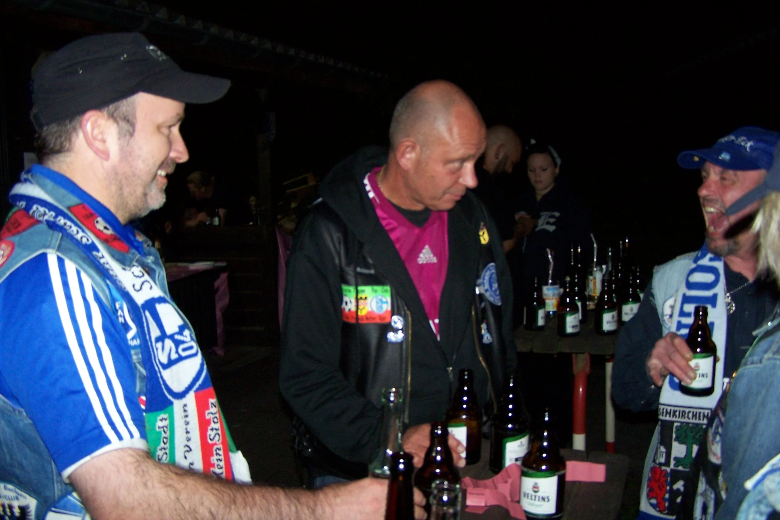Drei Männer in Sportkleidung und Zipfelmützen, die Bierflaschen in der Hand halten, beim gemeinsamen Trinken und Unterhalten in einer dunklen Bar oder einem Club.