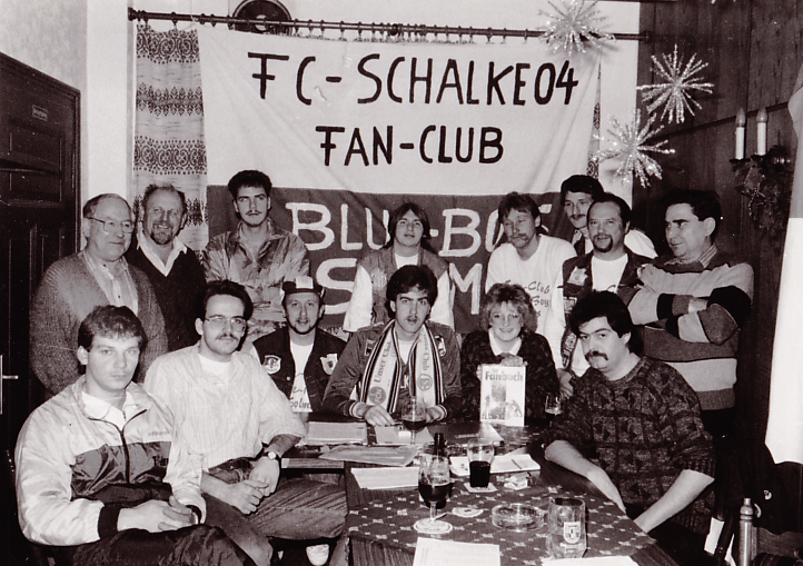Gruppe von Menschen, die bei einer FC Schalke 04 Fan-Club-Veranstaltung an einem Tisch sitzen, mit Bannern im Hintergrund, die den Fan-Club anzeigen.