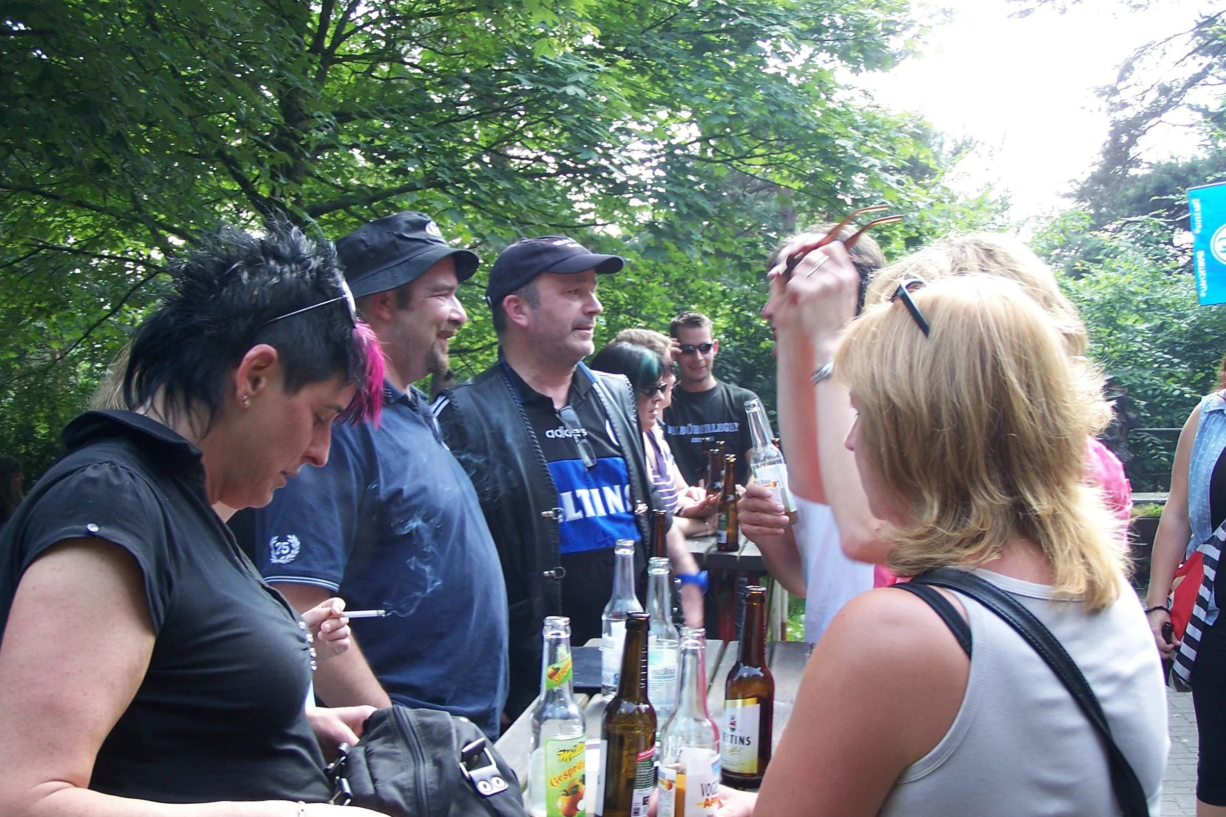 Menschen bei einem Treffen im Freien, einige halten Getränke, im Hintergrund grüne Bäume, sonniges Wetter.