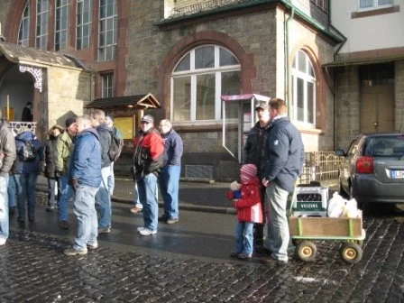 Menschen stehen in einer Schlange auf einer nassen Straßenfläche vor einem alten Backsteingebäude mit großen Fenstern. Ein Kind mit rotem Mantel zieht einen kleinen Wagen.