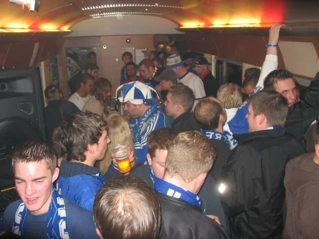 Menschen feiern in einem Bus, viele tragen Schal oder Kleidung in den Farben Blau und Weiß, einige halten Bier in der Hand.